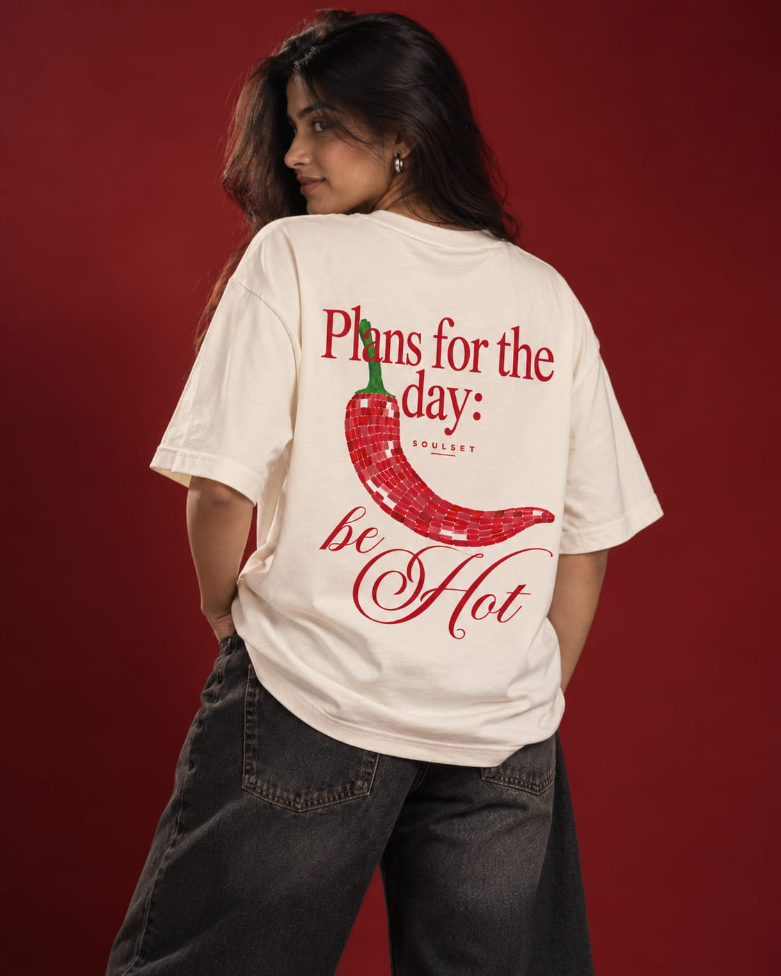 A woman wearing a cream-colored graphic t-shirt with a red chili pepper design and the text 'Plans for the day: be Hot' on the back, paired with dark gray jeans, set against a solid dark red background.