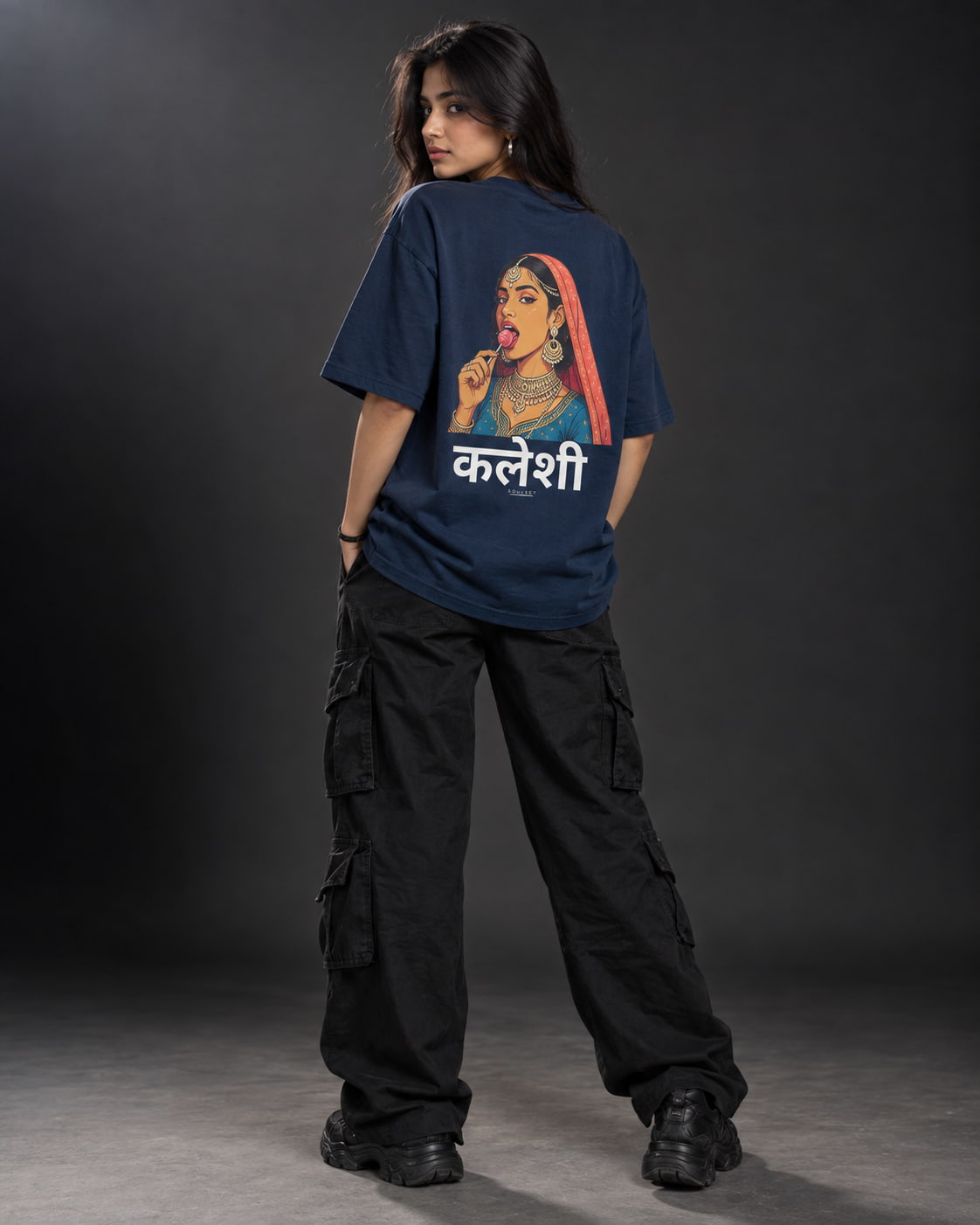 A woman stands with her back to the camera wearing a navy blue oversized graphic t-shirt featuring a colorful illustration of a woman in a red headscarf and white Devanagari text, paired with black cargo pants and black shoes, against a dark studio background.