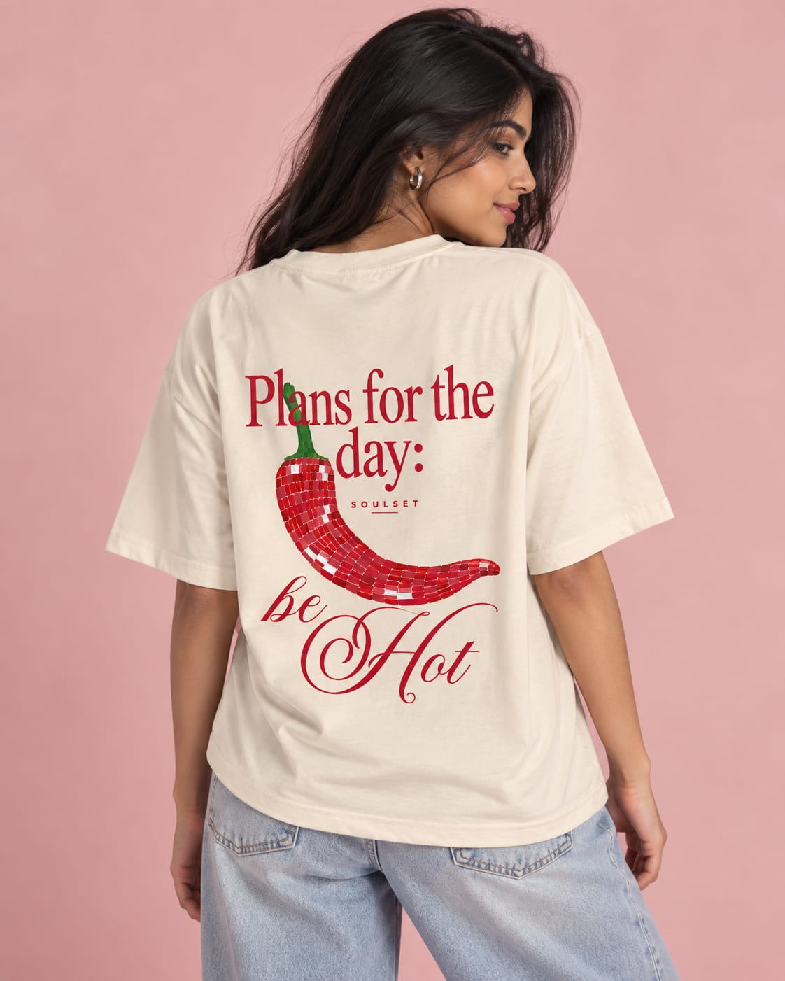 A woman wearing an oversized cream-colored t-shirt with a red chili pepper graphic and the text 'Plans for the day: be Hot' on the back, paired with light blue jeans against a solid pink background.
