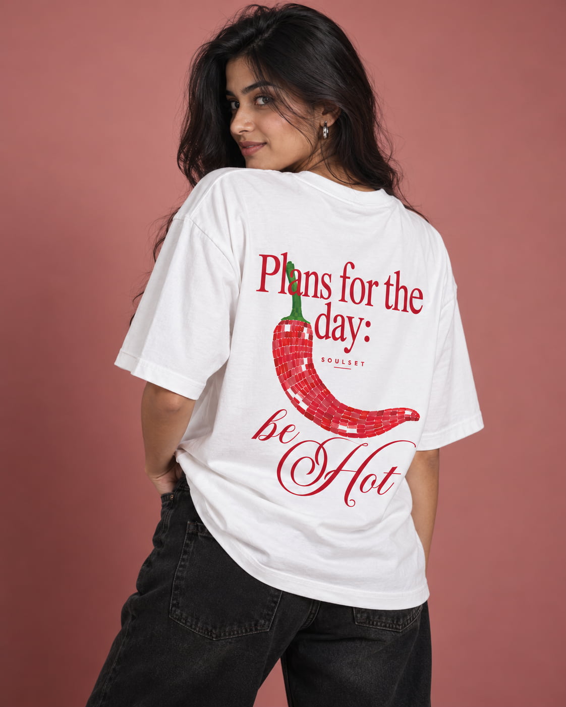 A woman wearing a white graphic t-shirt with a red chili pepper design and the text 'Plans for the day: be Hot' on the back, paired with black jeans against a dusty rose background.