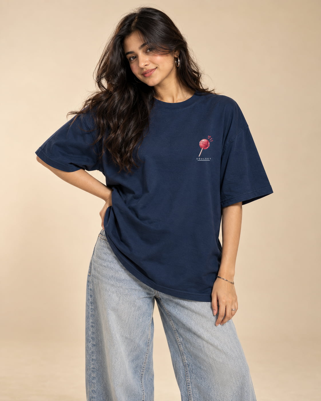 A woman modeling an oversized navy blue t-shirt with a small pink and white lollipop graphic on the left chest, paired with light-wash wide-leg jeans, against a solid beige studio background.