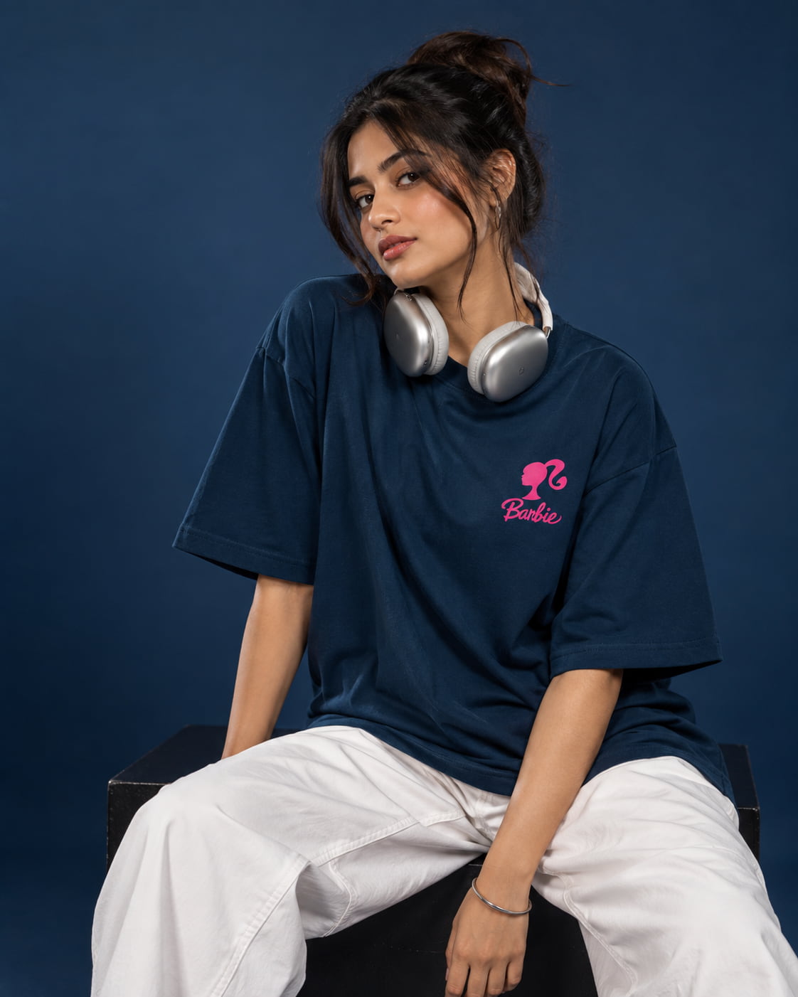 A woman wearing an oversized navy blue t-shirt with a pink Barbie logo on the chest, paired with white pants and silver headphones around her neck, sitting against a dark blue background.