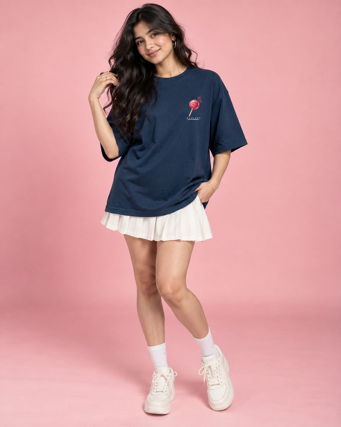 A woman modeling an oversized navy blue t-shirt with a small pink lollipop graphic on the left chest, paired with a white pleated mini skirt, white socks, and white sneakers, set against a solid pink studio background.