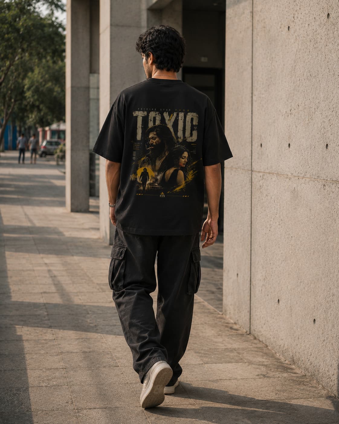 A man walking away on a concrete sidewalk, wearing a black graphic t-shirt with a large back print that reads 'TOXIC' and 'BREATHE' with a yellow and dark illustration, paired with dark cargo pants and white sneakers.