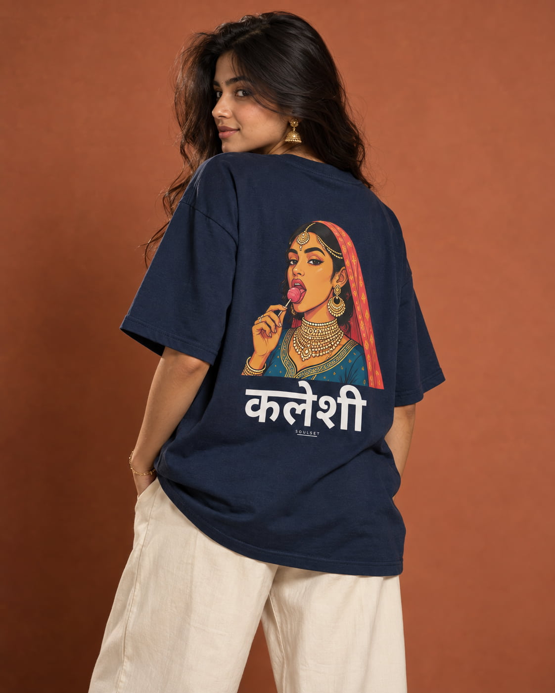 A woman wearing an oversized navy blue graphic T-shirt with a stylized illustration of a woman in traditional Indian attire and white Hindi text on the back, paired with cream-colored pants, set against a solid terracotta background.