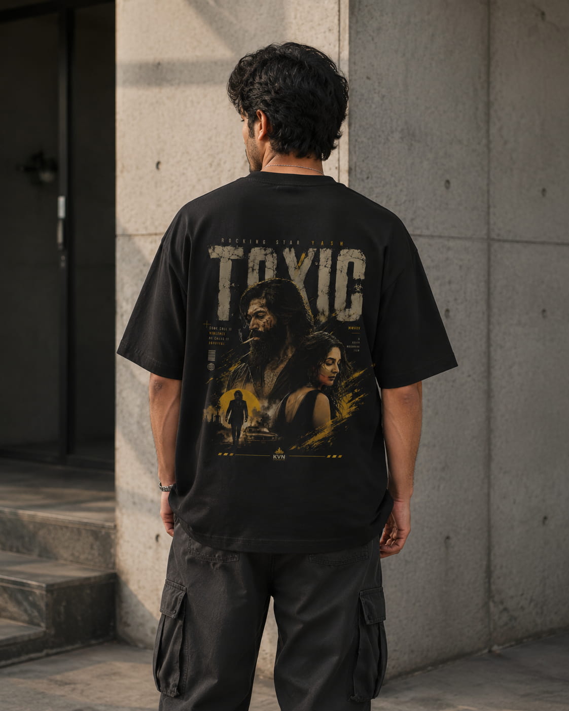 A man viewed from behind wearing a black graphic t-shirt with a large distressed print reading 'TOXIC' and a dark scene, paired with dark cargo pants, standing against a concrete wall.
