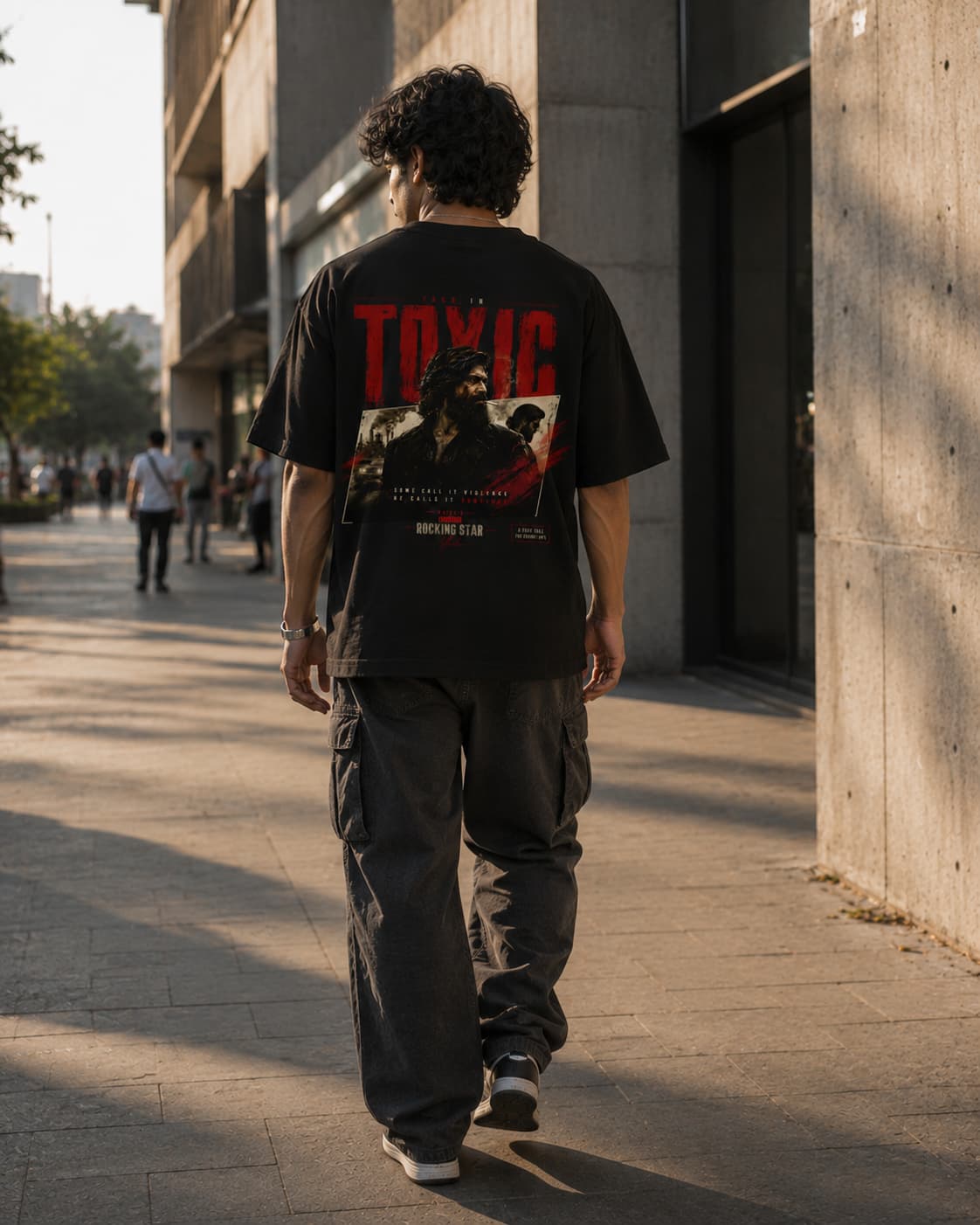 A man viewed from behind walking on a city sidewalk, wearing a black graphic T-shirt with a large red and white 'TOXIC' print and dark cargo pants.