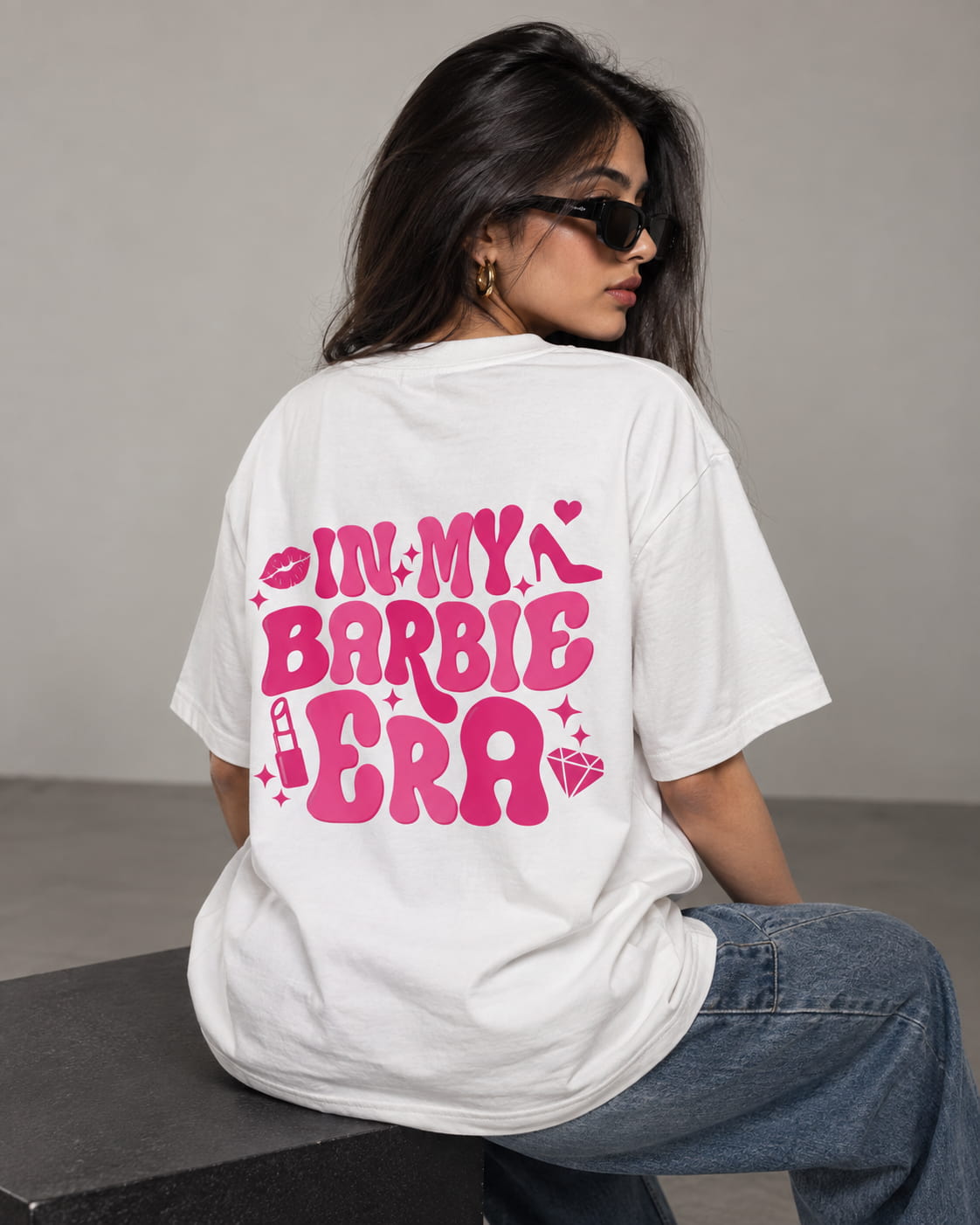 A woman wearing a white oversized T-shirt with a bold pink graphic on the back that reads 'IN MY BARBIE ERA' with small decorative icons, seated against a plain gray wall.