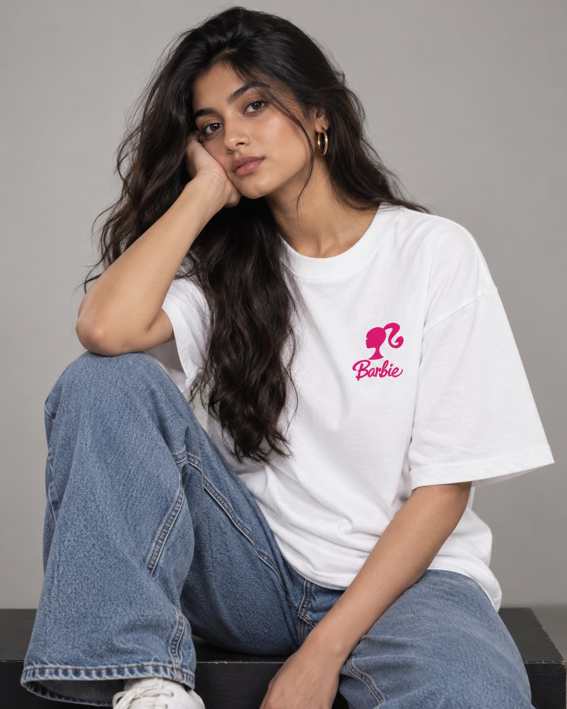 A woman sitting and wearing a white short-sleeve T-shirt with a pink Barbie logo on the left chest, paired with wide-leg blue jeans against a plain gray studio background.