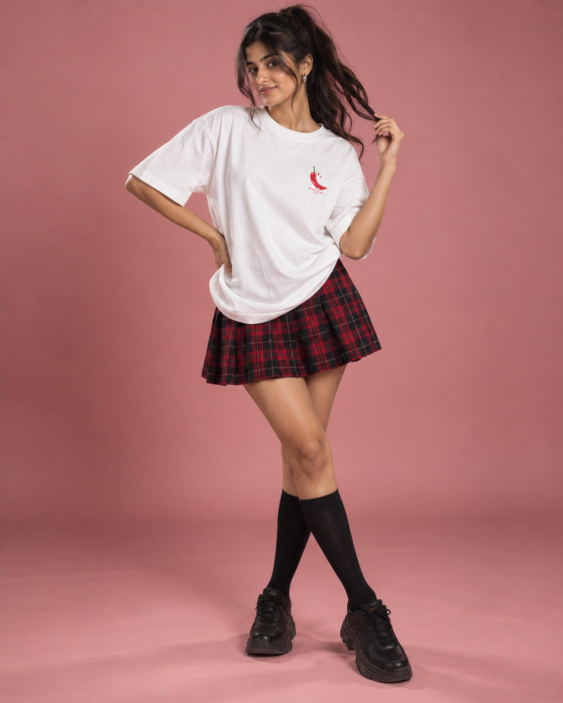 A woman modeling a white oversized t-shirt with a small red chili pepper graphic on the left chest, paired with a red and black plaid pleated mini skirt, black knee-high socks, and black platform sneakers, set against a solid pink studio backdrop.