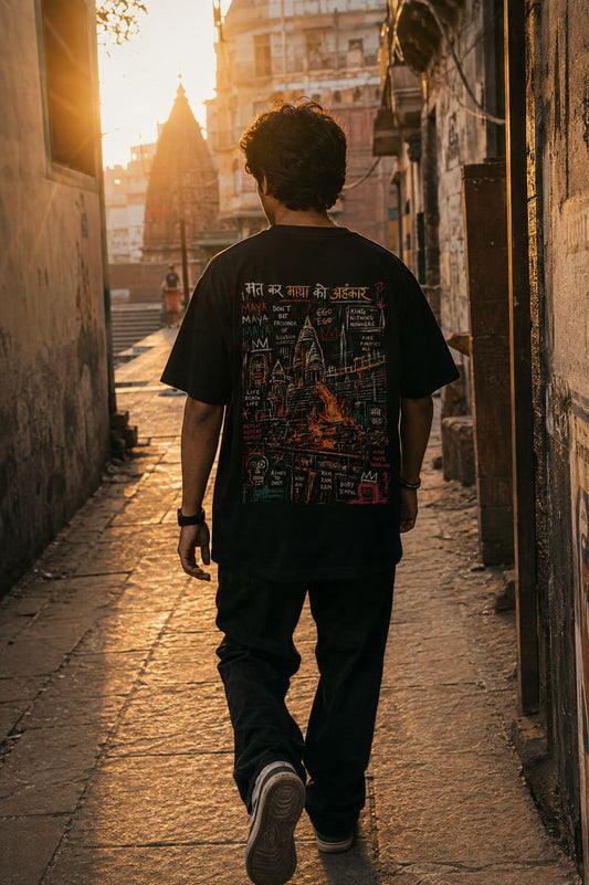 A man viewed from behind walking down a sunlit stone alleyway, wearing a black graphic T-shirt with a large multicolor back print and dark pants.