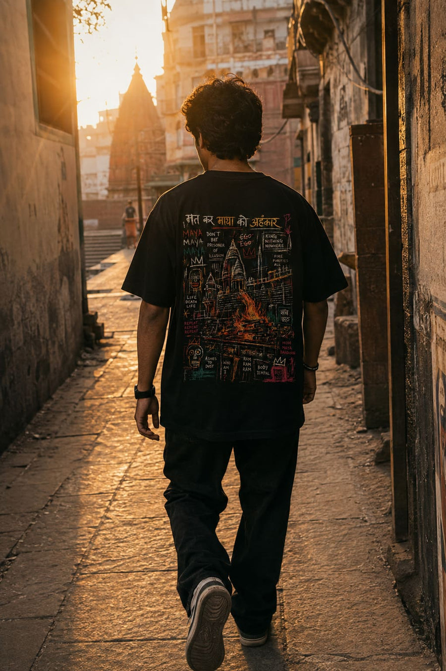 A man viewed from behind walking down a sunlit stone alleyway, wearing a black graphic T-shirt with a large multicolor back print and dark pants.